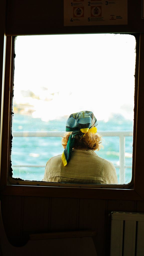 A woman with a headscarf gazes out a window at the sea, evoking serenity.
